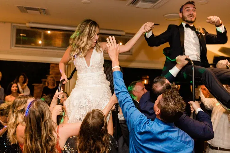 Wedding guests lift a smiling bride and groom on a dance floor, arms raised in celebration.