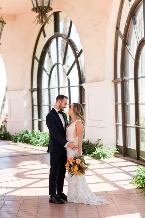 Bride and groom standing together in a sunlit arched hallway, holding hands and facing each other