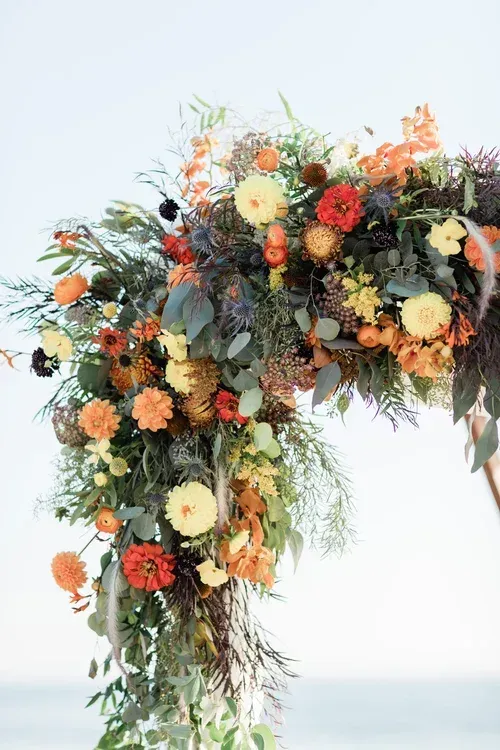 Floral arch of orange, cream, and green flowers against a pale sky and sea backdrop