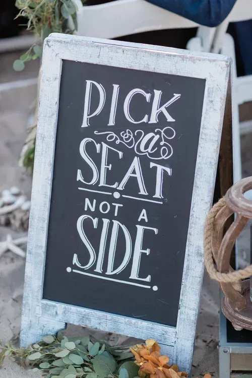 White rustic signboard with chalk lettering: “PICK a SEAT not a SIDE.”