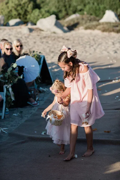 Two children in pastel dresses walk barefoot on a beach path carrying a basket and bag