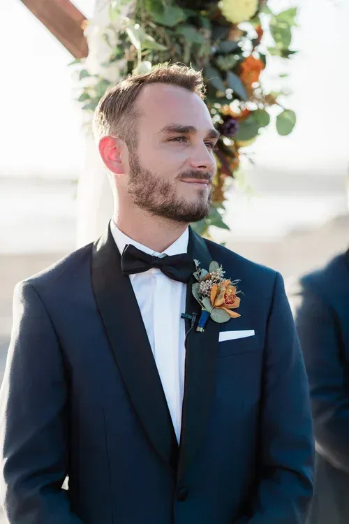Man in a navy tuxedo with floral boutonniere at an outdoor wedding ceremony