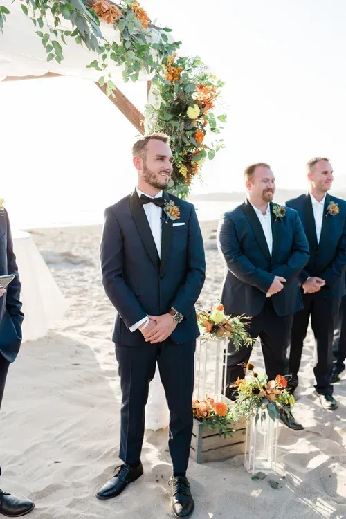 Three suited groomsmen stand under a floral arch on a sandy beach during a wedding ceremony
