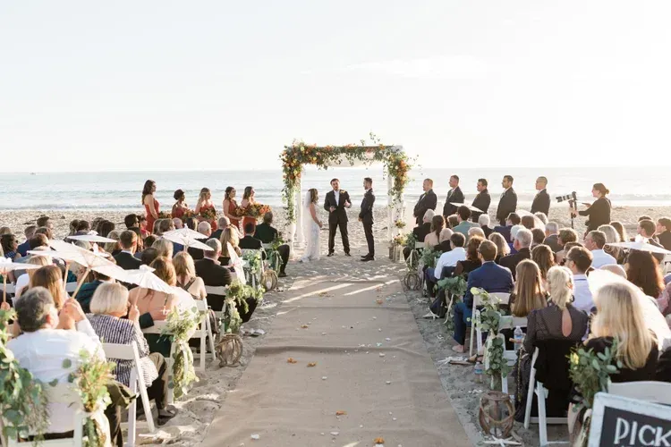 Outdoor beach wedding ceremony with guests seated along an aisle under a floral arch at sunset