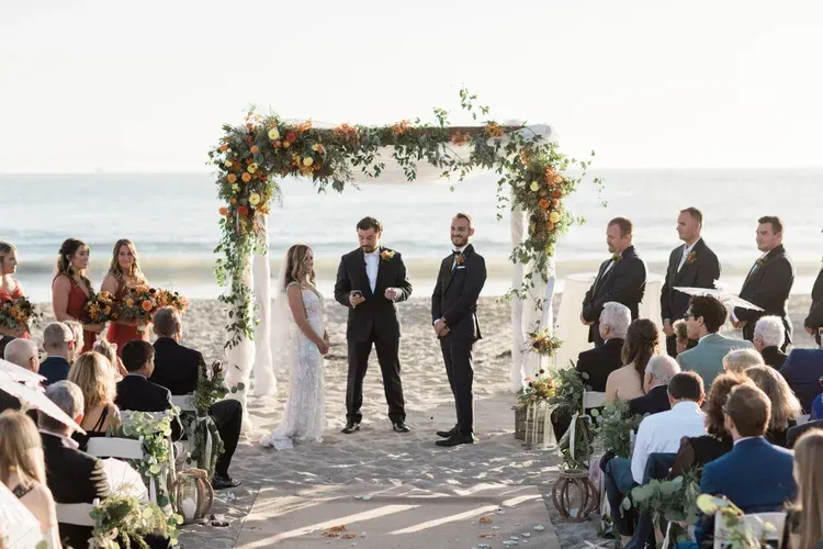 Wedding ceremony under a floral arch beside the ocean, with guests seated on both sides.
