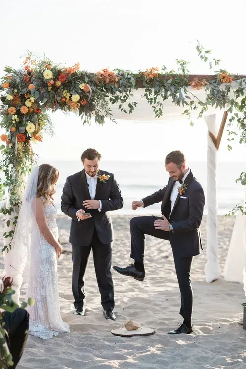 Wedding ceremony under floral arch; two grooms and a bride stand on a sandy aisle.