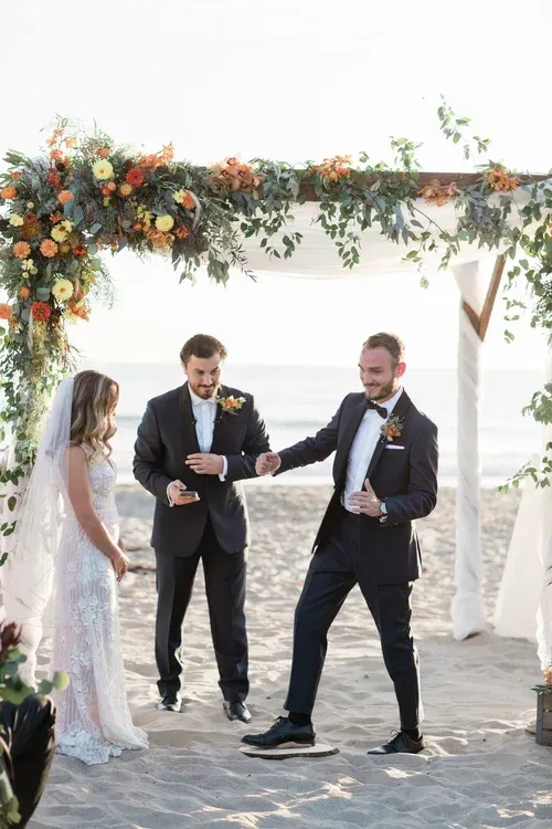 Wedding ceremony under floral arch, with bride and two grooms standing on a stone path outdoors.