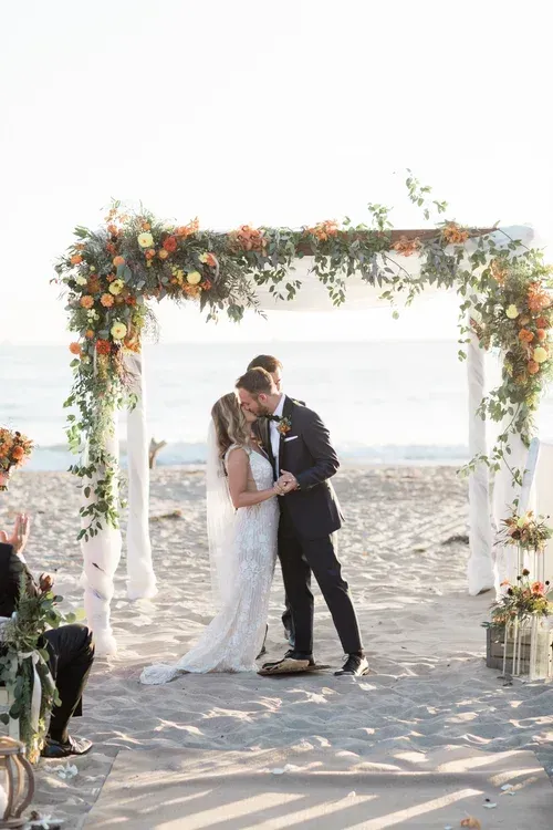Bride and groom kissing beneath a flower-covered arch on a beach ceremony setting