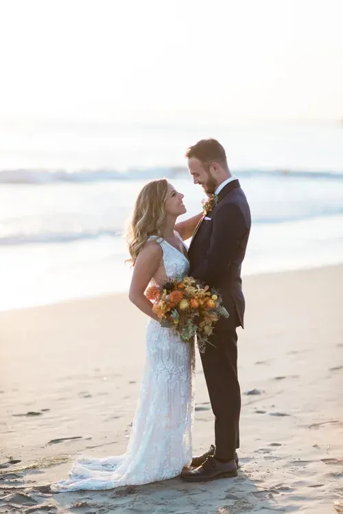 Couple in wedding attire standing on a beach, holding a bouquet at sunset