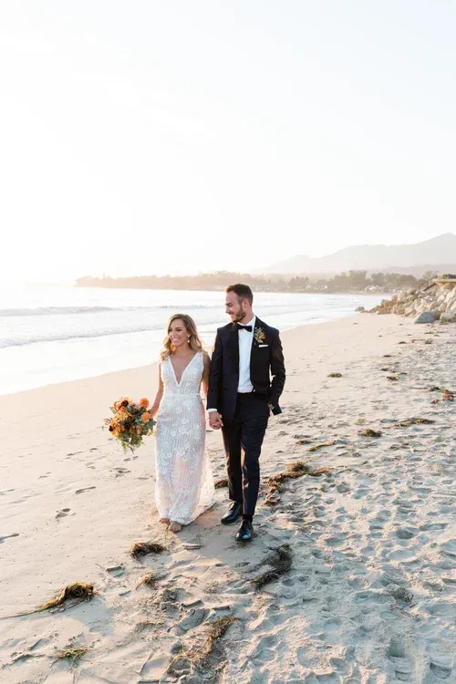 Couple walking along a sunny beach, bride in white dress holding bouquet, groom in black tuxedo