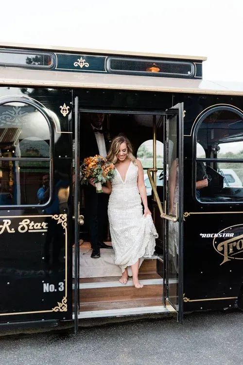Bride in white dress steps down from a vintage black trolley with floral arrangements.