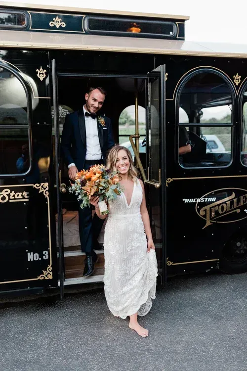 Bride in lace dress steps from a black trolley while holding a bouquet; groom stands in the doorway.