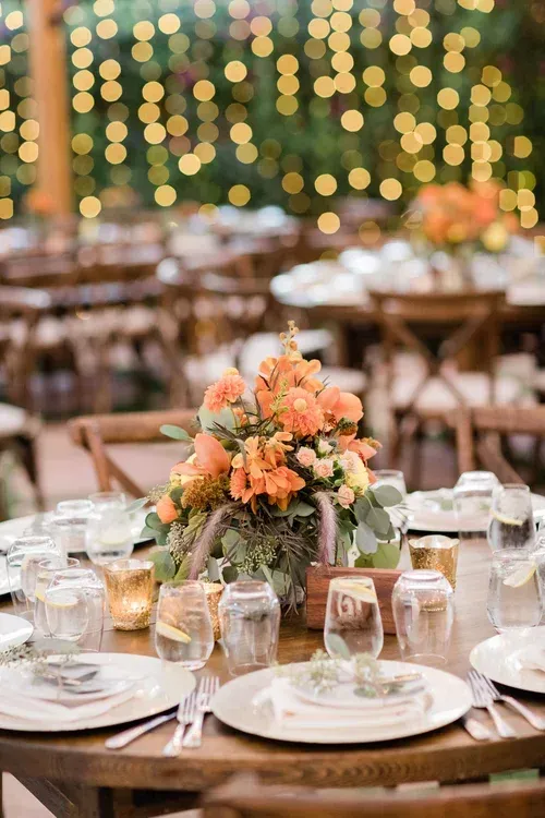 Floral centerpiece on a wooden wedding reception table with plates, glasses, and warm string lights in the background