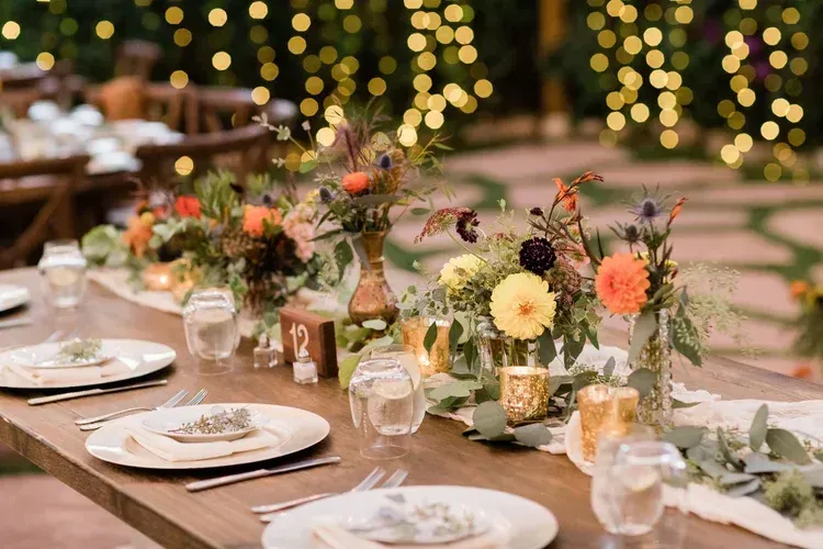 Elegant outdoor table setting with white plates, floral centerpieces, and warm string lights bokeh
