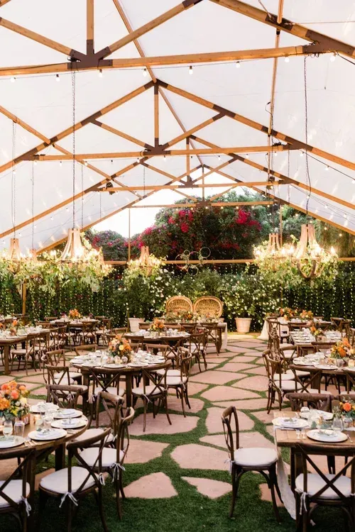 Outdoor wedding reception under a transparent tent with wooden beams, round tables, chairs, and floral decor