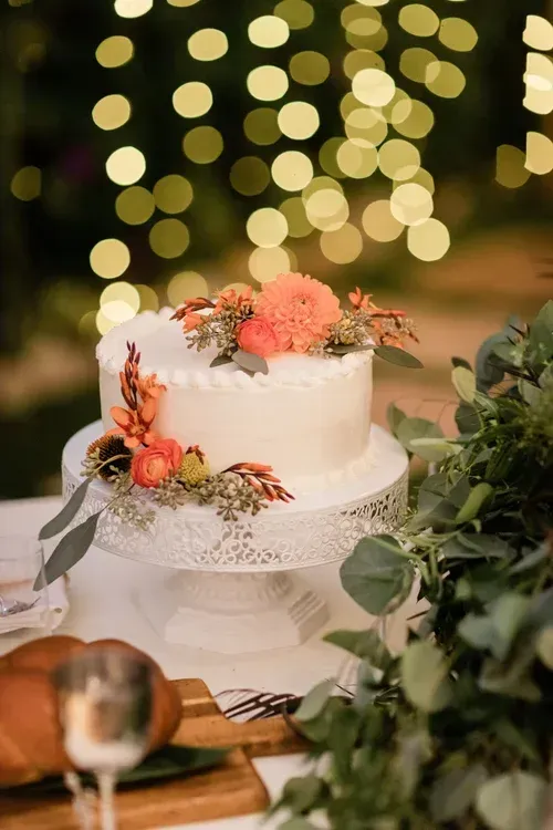 Floral-decorated white cake on a lace stand with warm bokeh lights in the background.