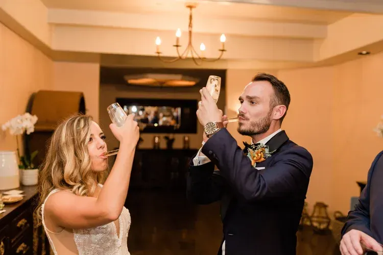 Two people in formal attire raising glasses during a toast in a warmly lit indoor reception hall
