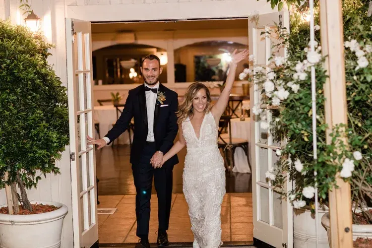 Newlyweds in formal attire smiling at a venue entrance with floral decorations