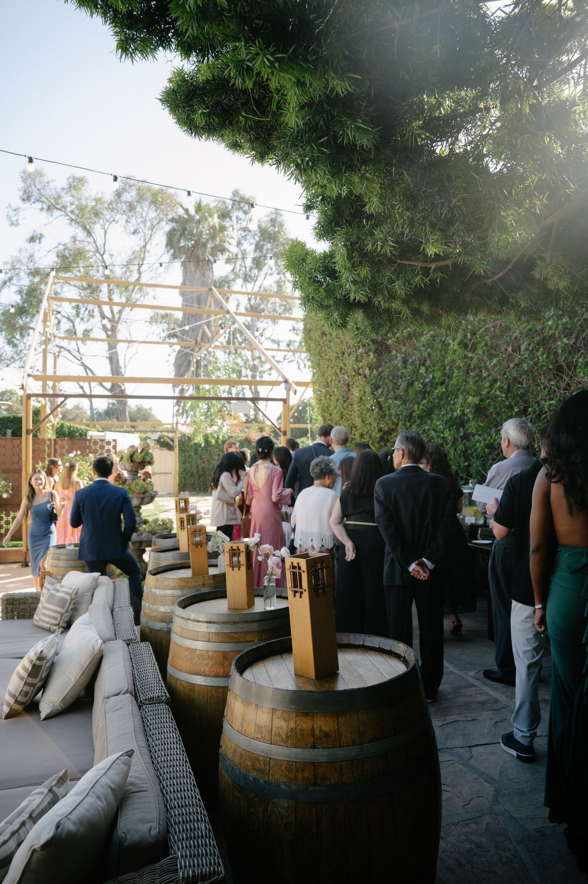 People gathered outdoors beside wooden barrels and hay bales under trees near a rustic structure.