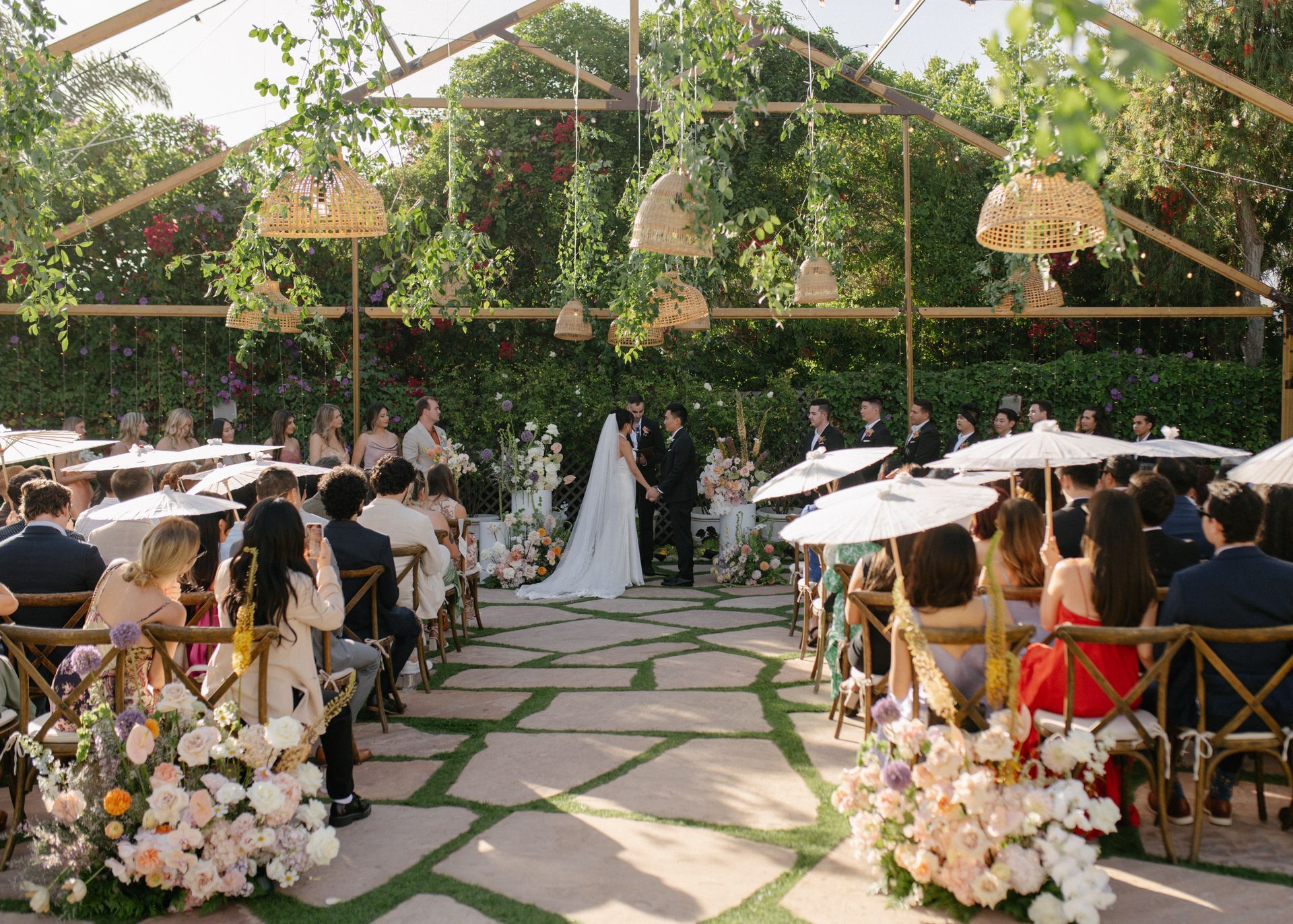 Outdoor wedding ceremony in a flower-filled garden with guests seated under white umbrellas and a couple at the altar