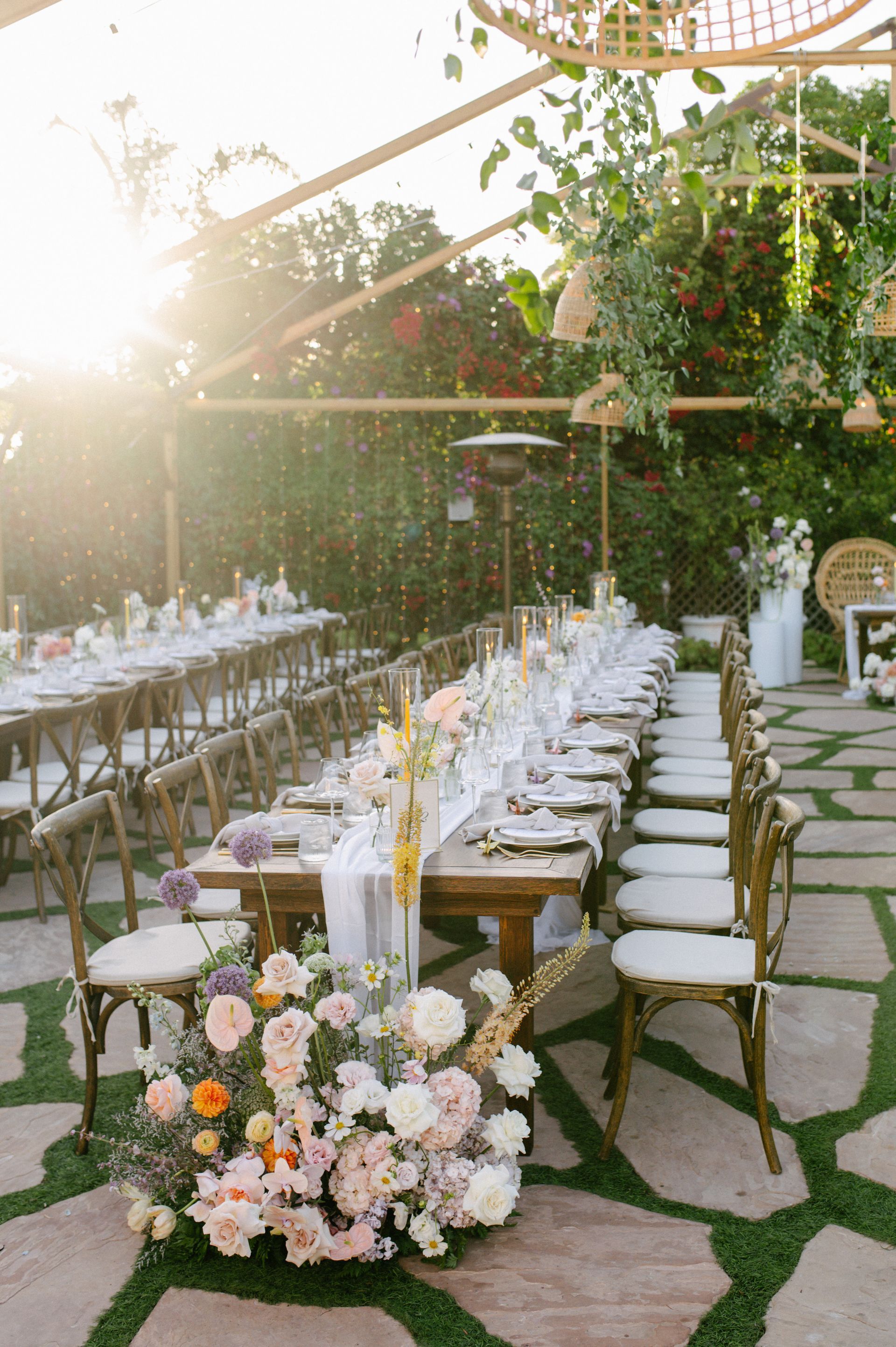Elegant outdoor reception with long flower-lined tables, white chairs, and greenery draped overhead at sunset