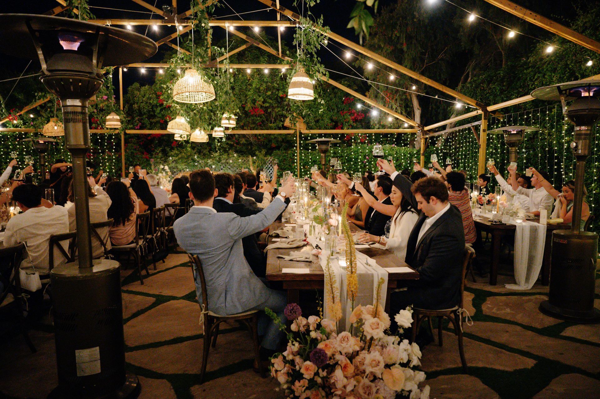 Guests toast at a candlelit outdoor wedding reception under string lights and greenery.