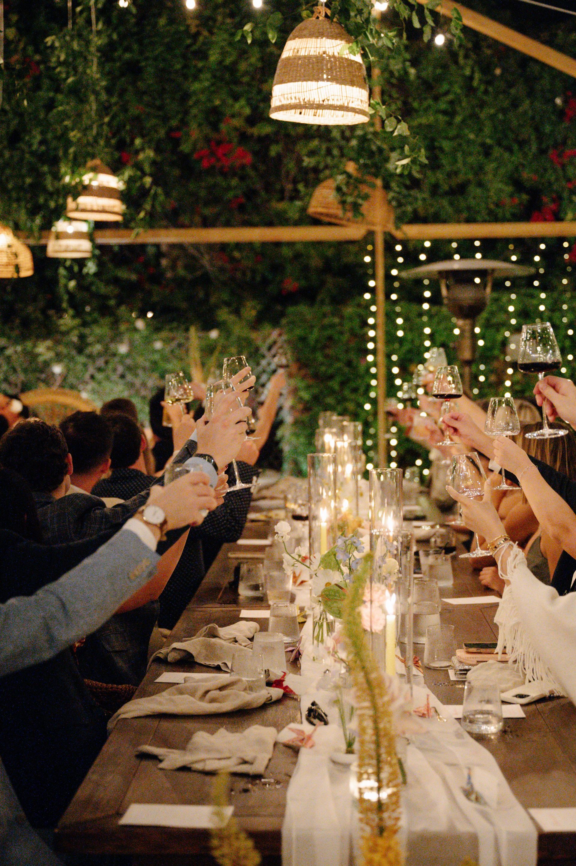 Guests raise glasses at a warmly lit outdoor dinner under string lights and greenery.