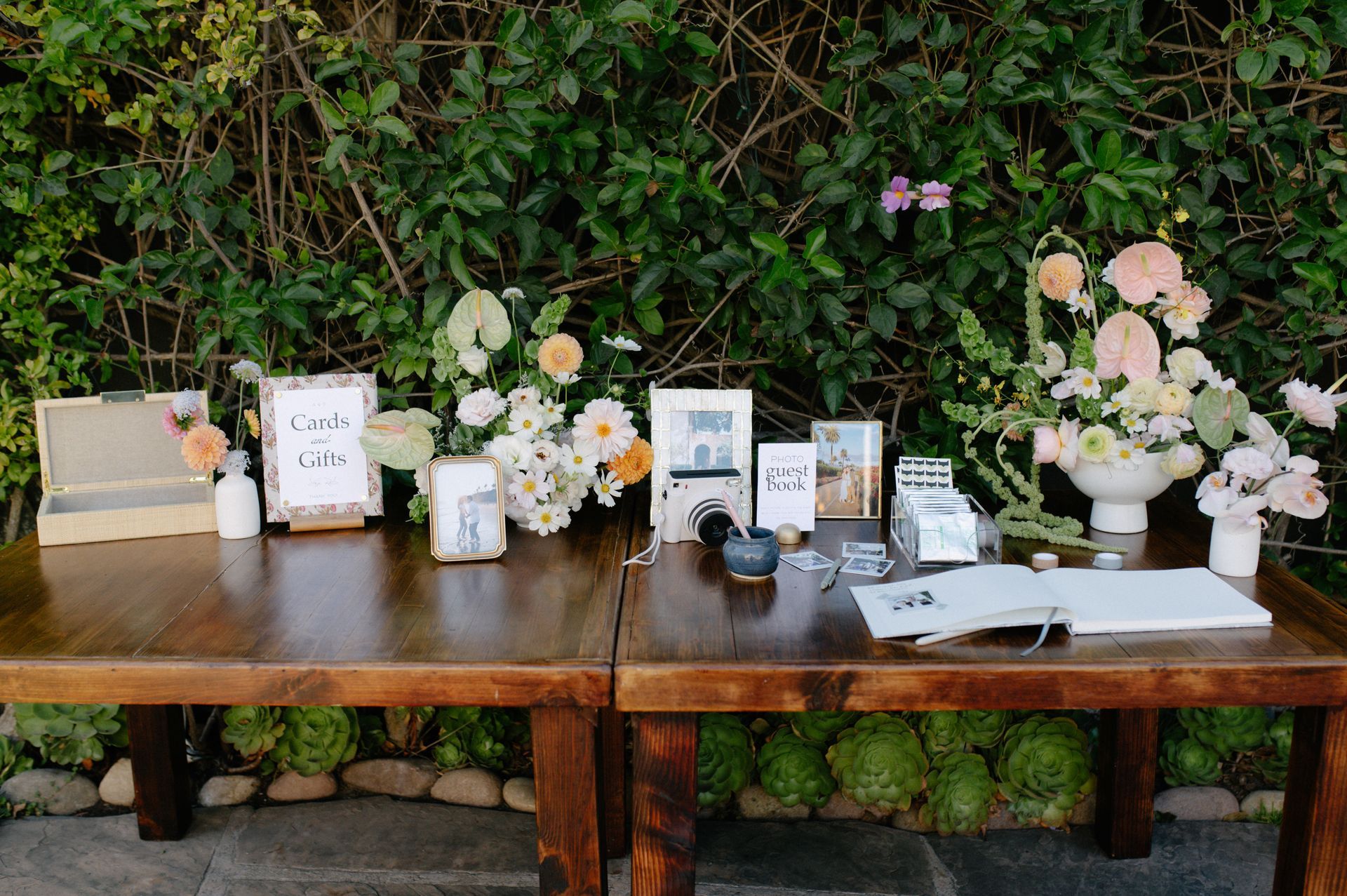 Two wooden tables with floral arrangements, framed signs, candles, and a guest book in a garden setting