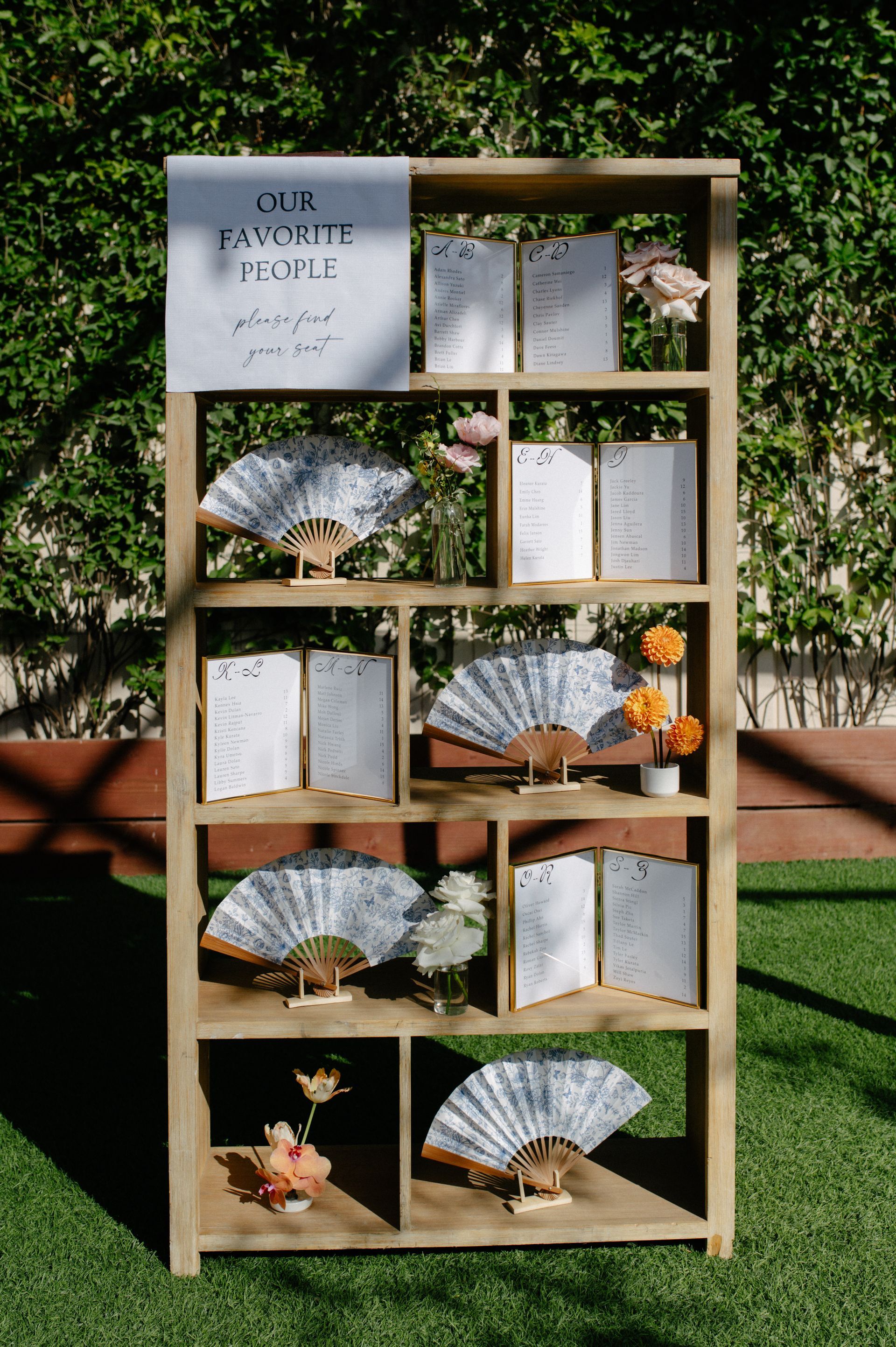 Decorative wooden shelf with open books and folded paper fans outdoors against a green hedge.