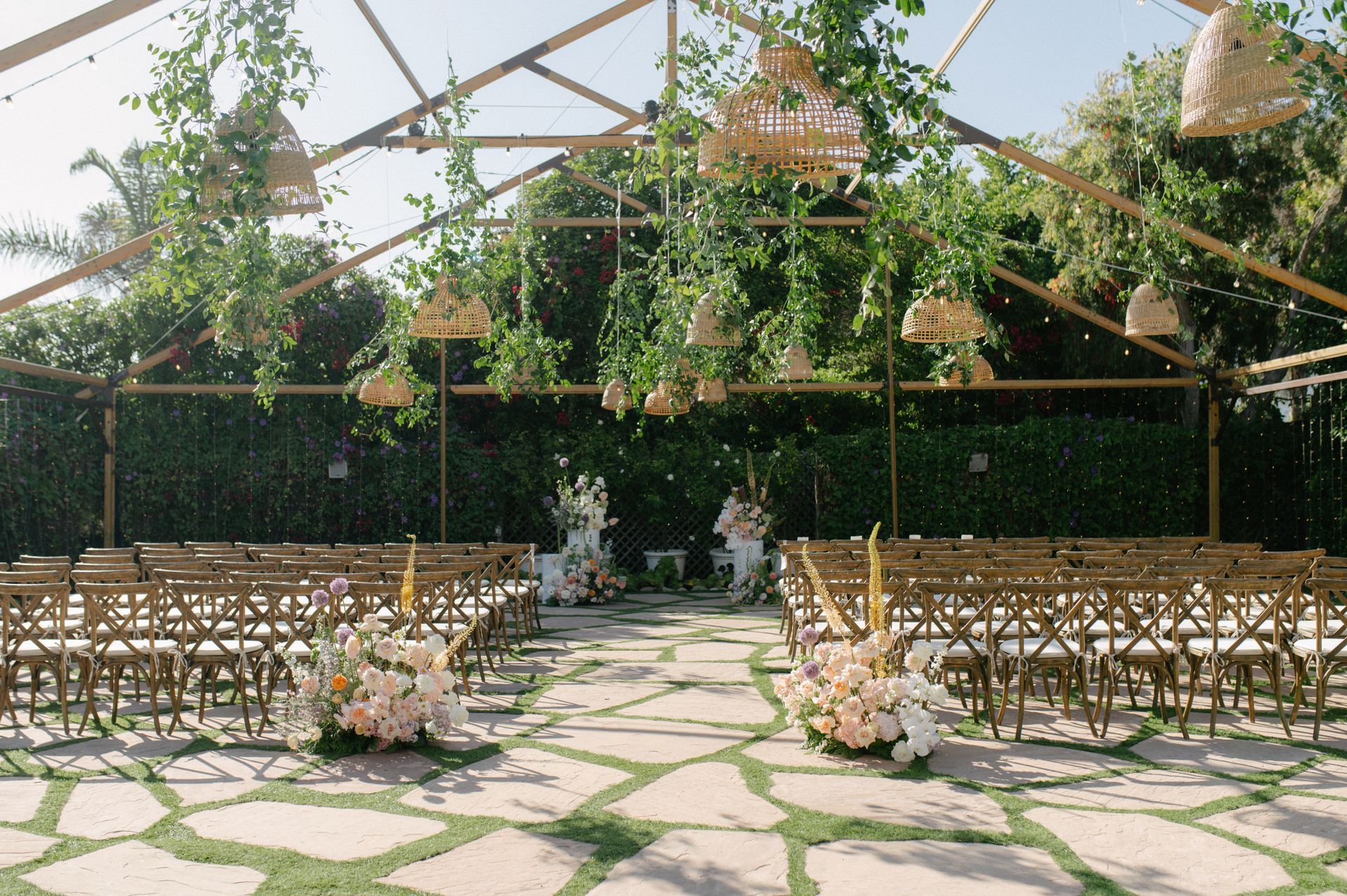 Outdoor wedding ceremony setup with rows of chairs, floral arrangements, and greenery under a glass canopy