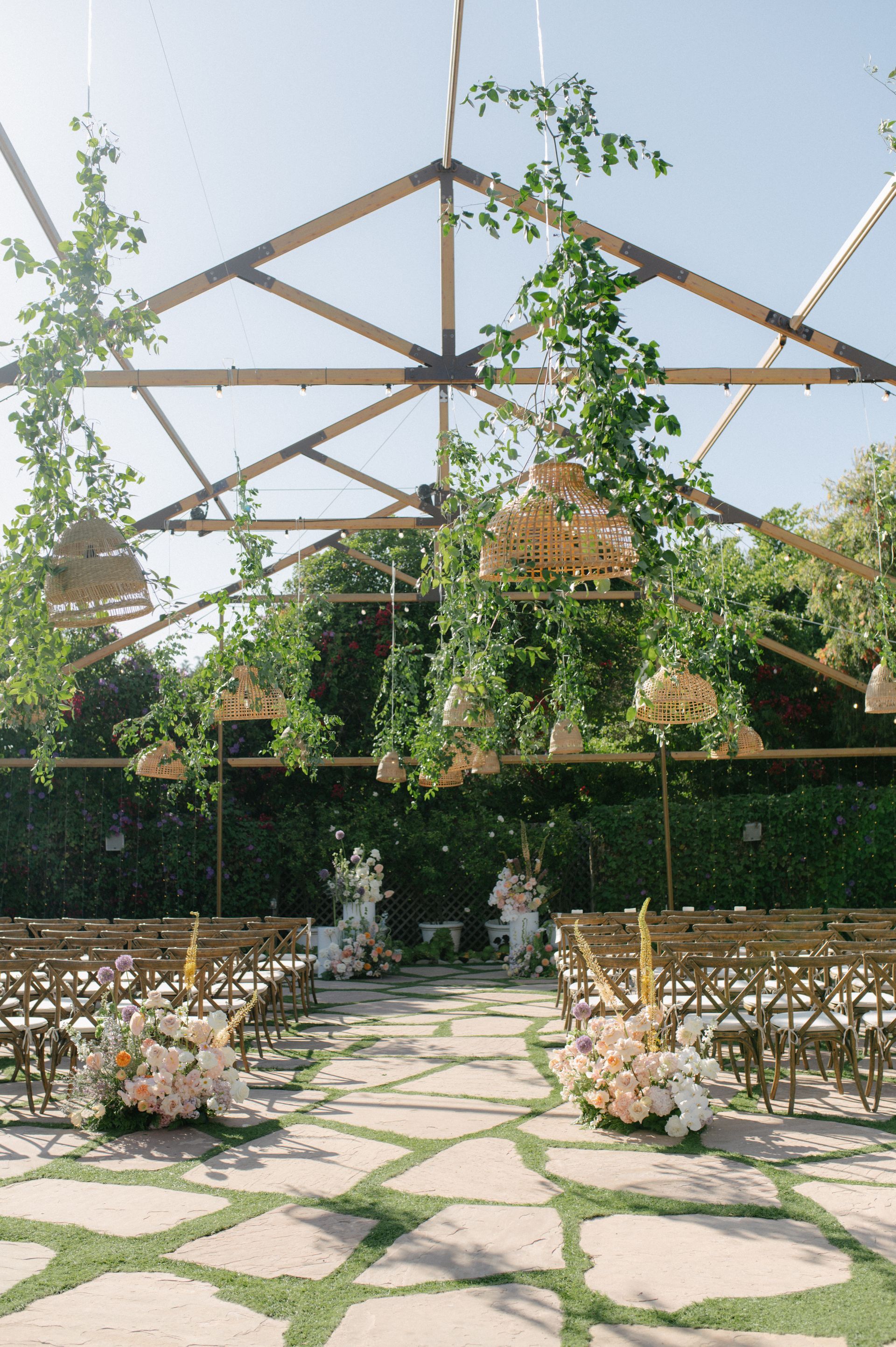 Outdoor wedding aisle with white chairs, floral arch, and a wooden canopy draped in greenery