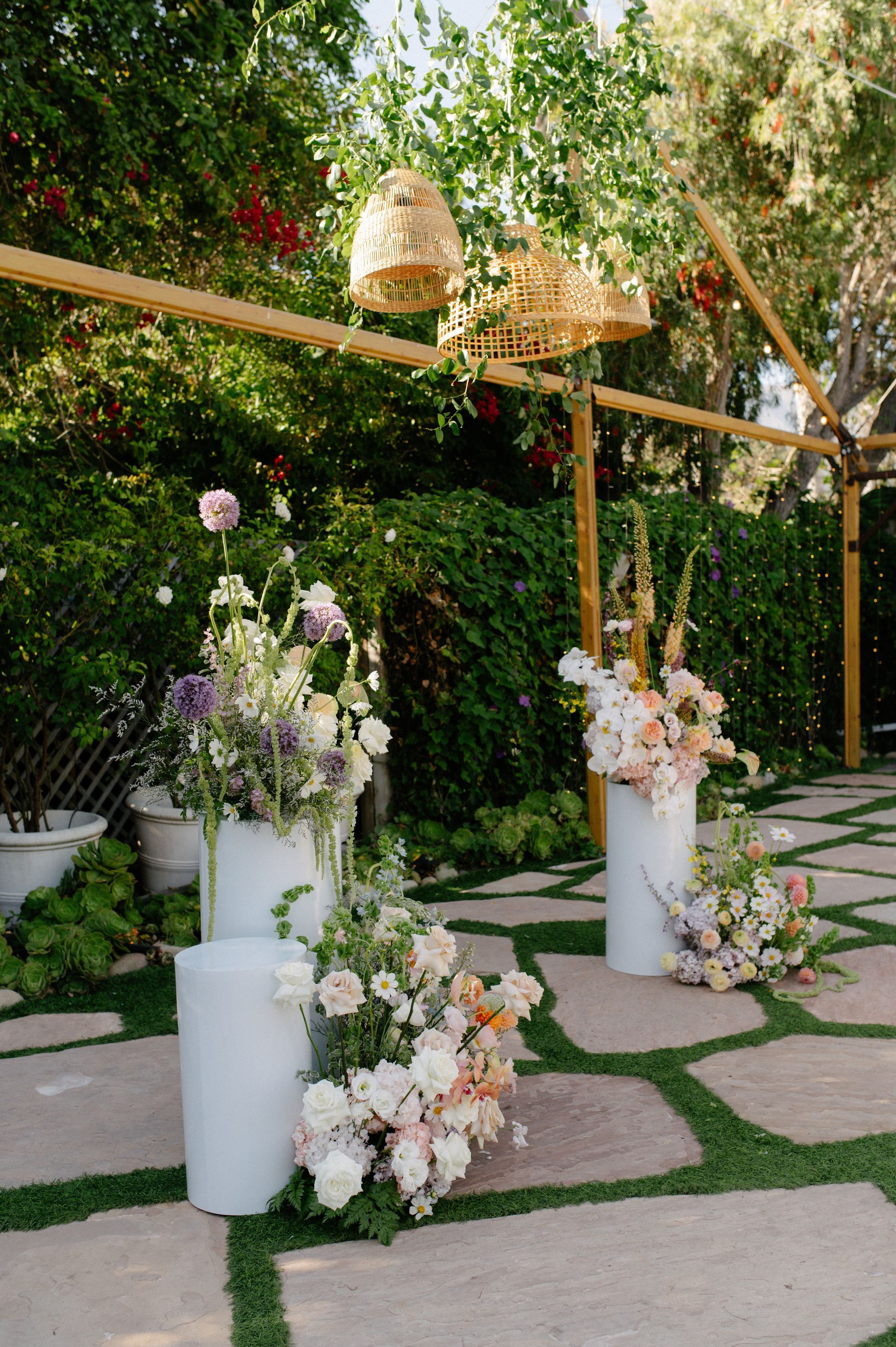 Floral outdoor ceremony setup with white pedestals, pastel arrangements, and hanging lanterns on a stone path