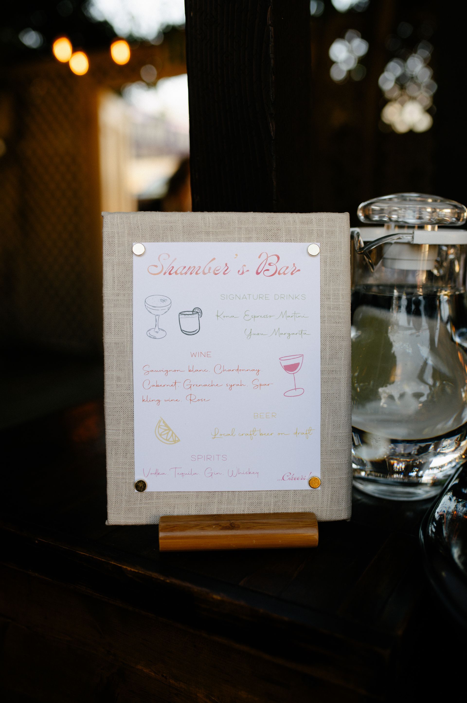 Small framed sign on a wooden stand beside a glass water pitcher on a dark table.
