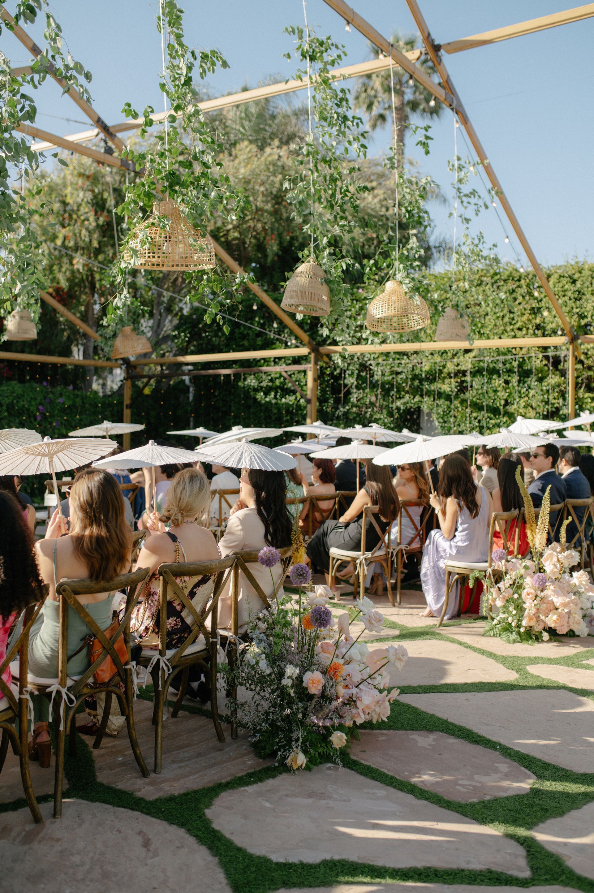 Outdoor wedding reception with guests at decorated tables under white umbrellas and greenery overhead
