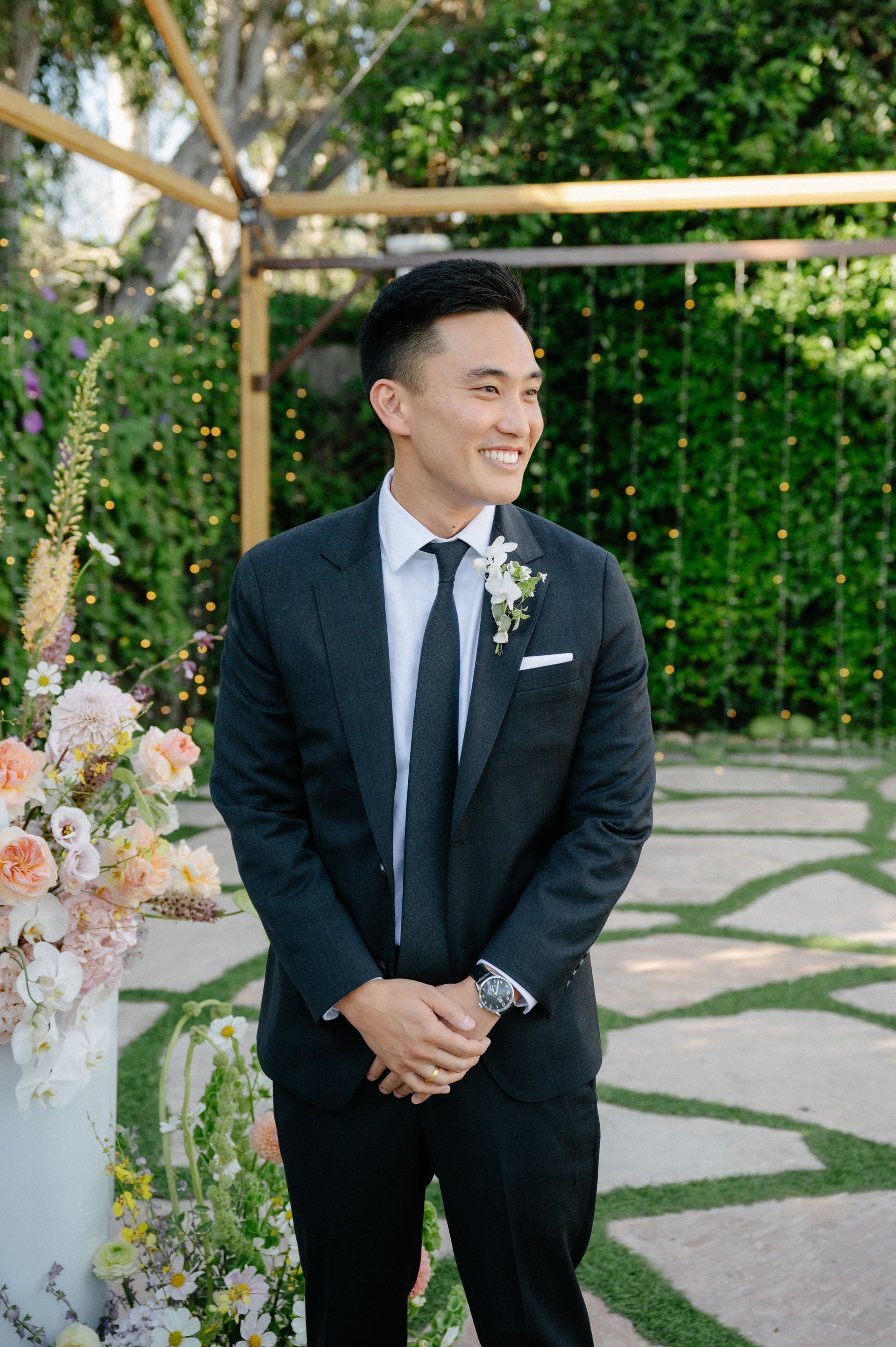 Man in a dark suit standing outdoors beside floral arrangements, smiling at a garden event