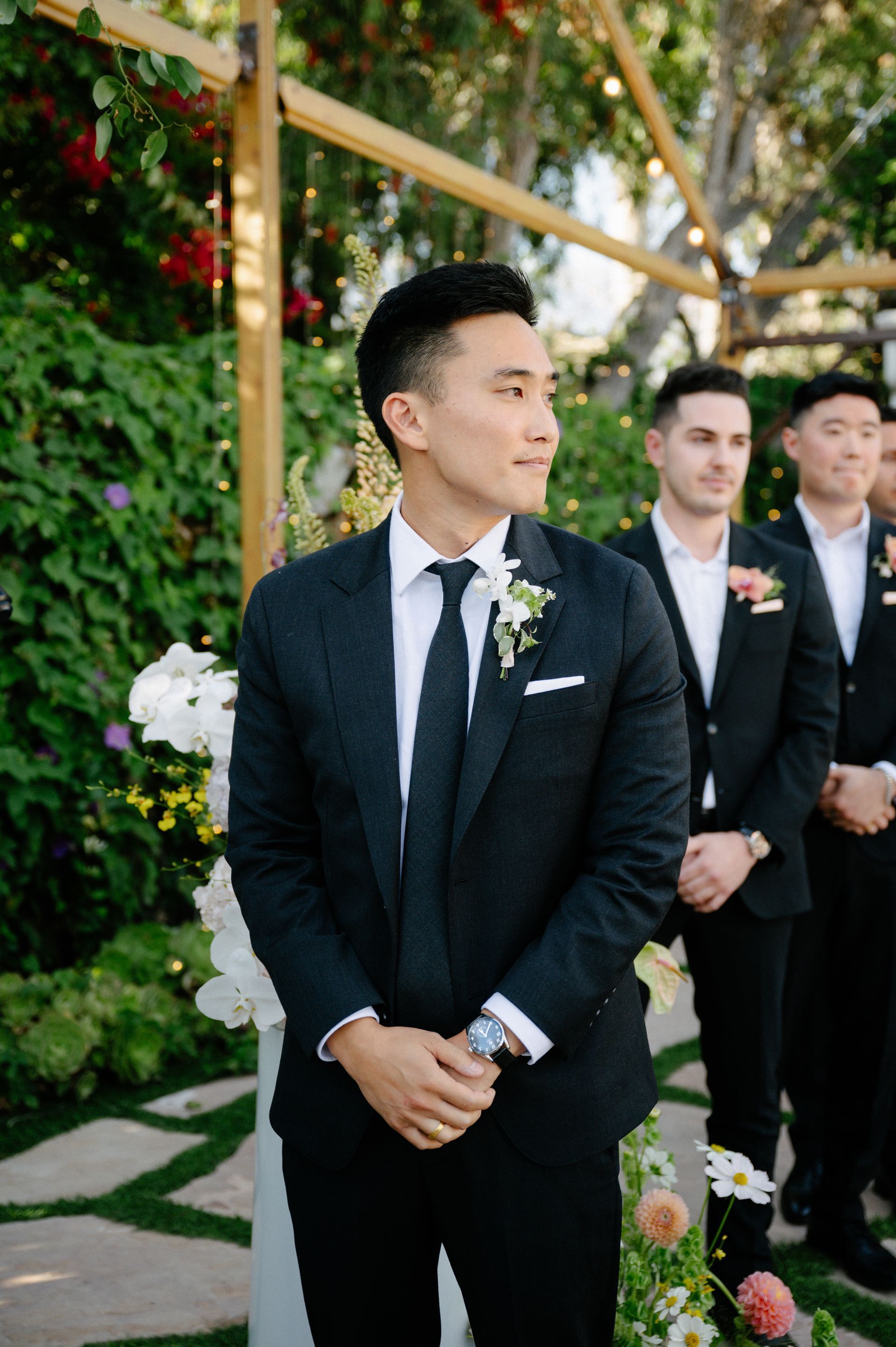 Man in a black suit and tie standing outdoors at a formal event with groomsmen behind him
