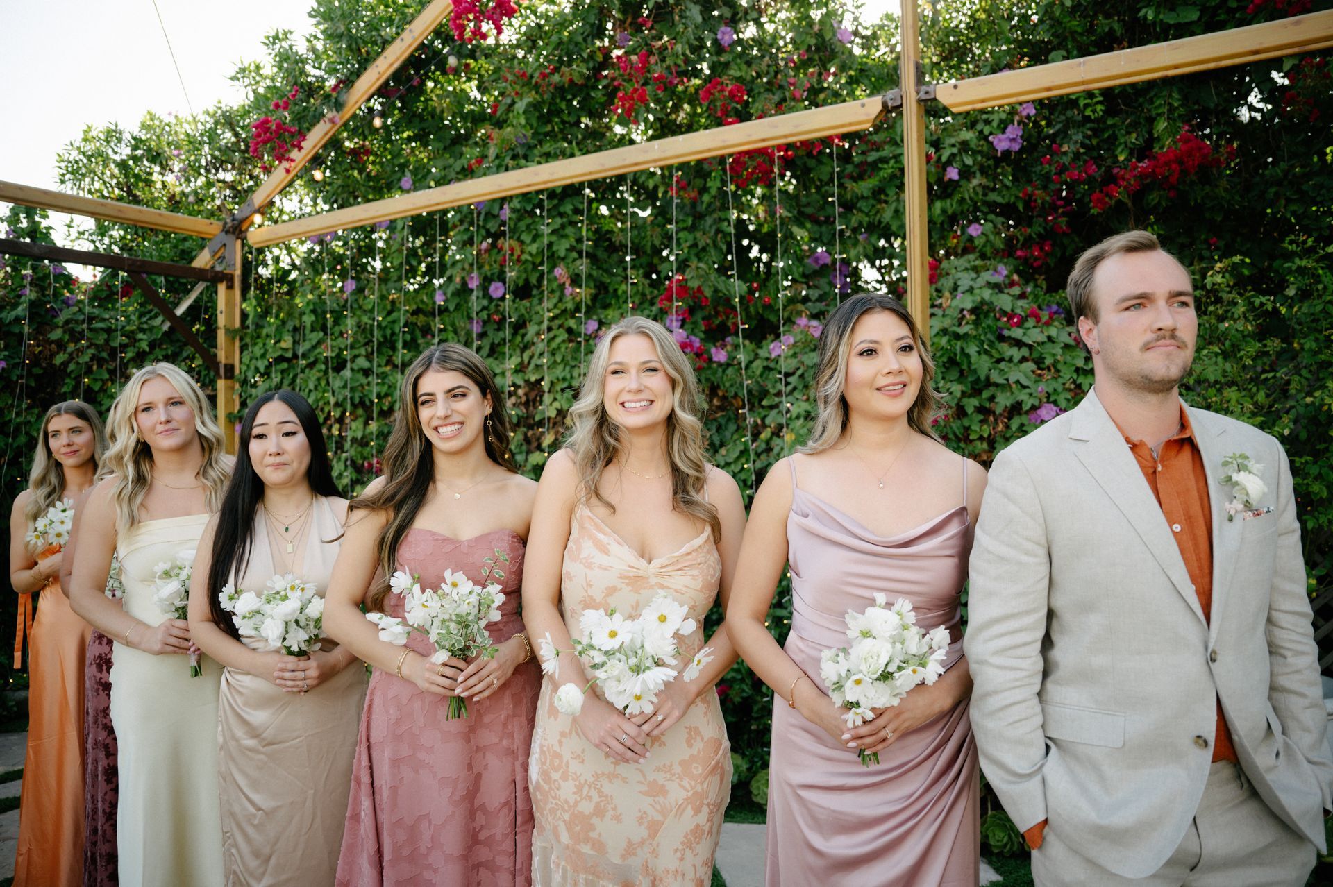 Wedding party in pink and cream dresses and a light suit, holding bouquets under a floral arbor outdoors