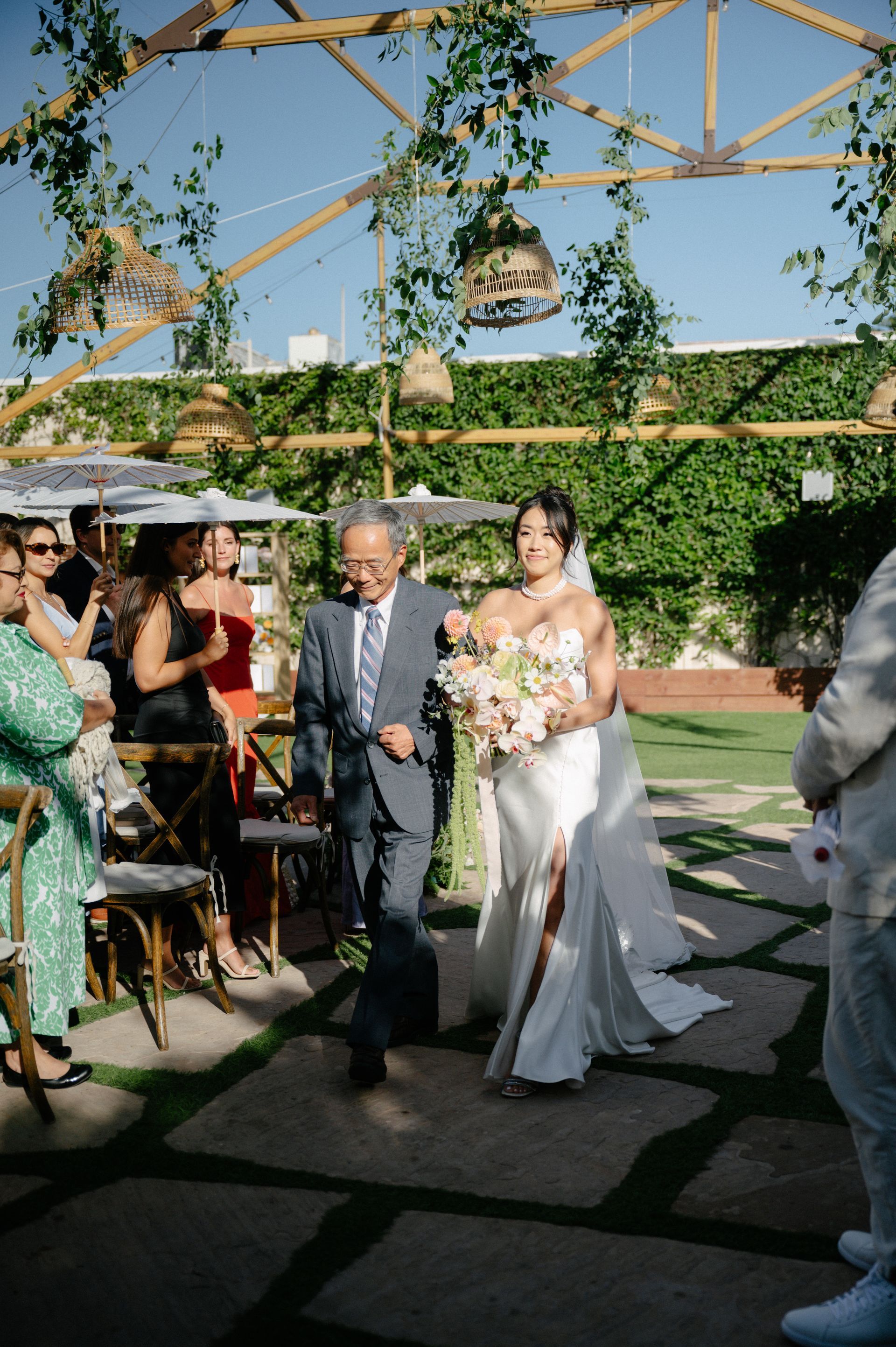 Bride in a white dress walking with an older man at an outdoor wedding reception under a vine-covered canopy