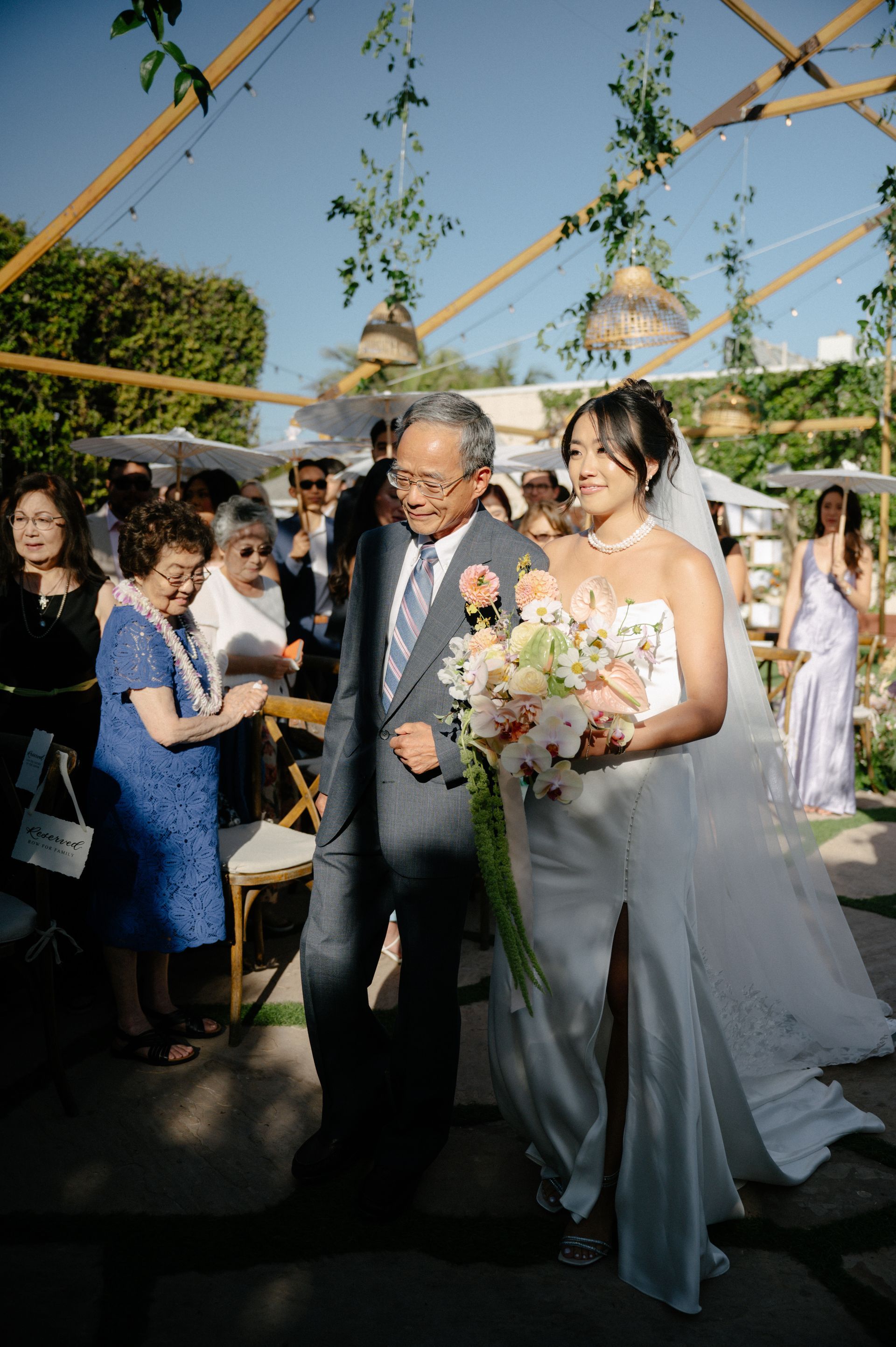 Bride and older man walking down outdoor wedding aisle, bride holding bouquet, guests nearby