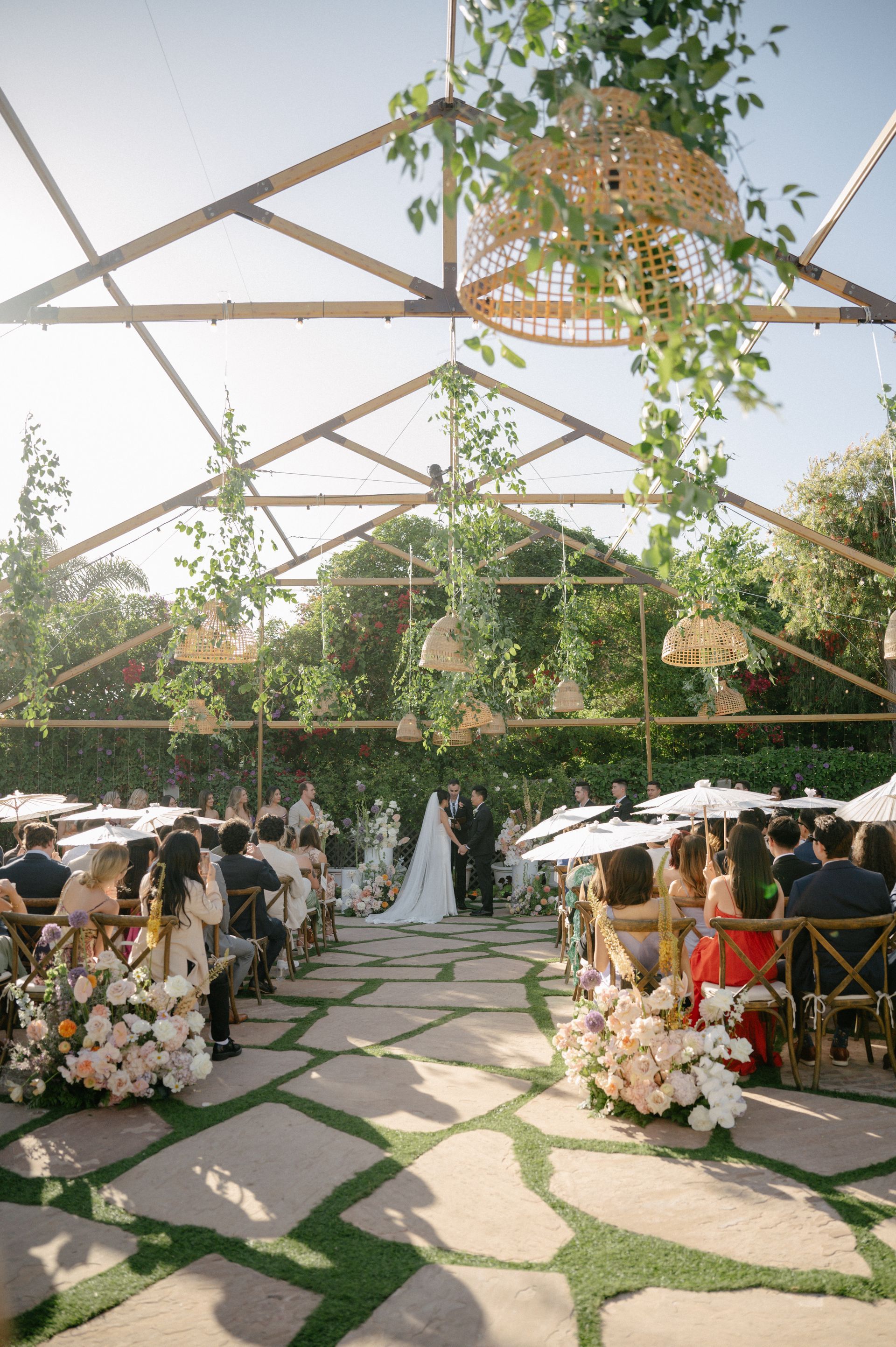 Outdoor wedding ceremony in a garden tent with white aisle flowers and seated guests under a geometric canopy
