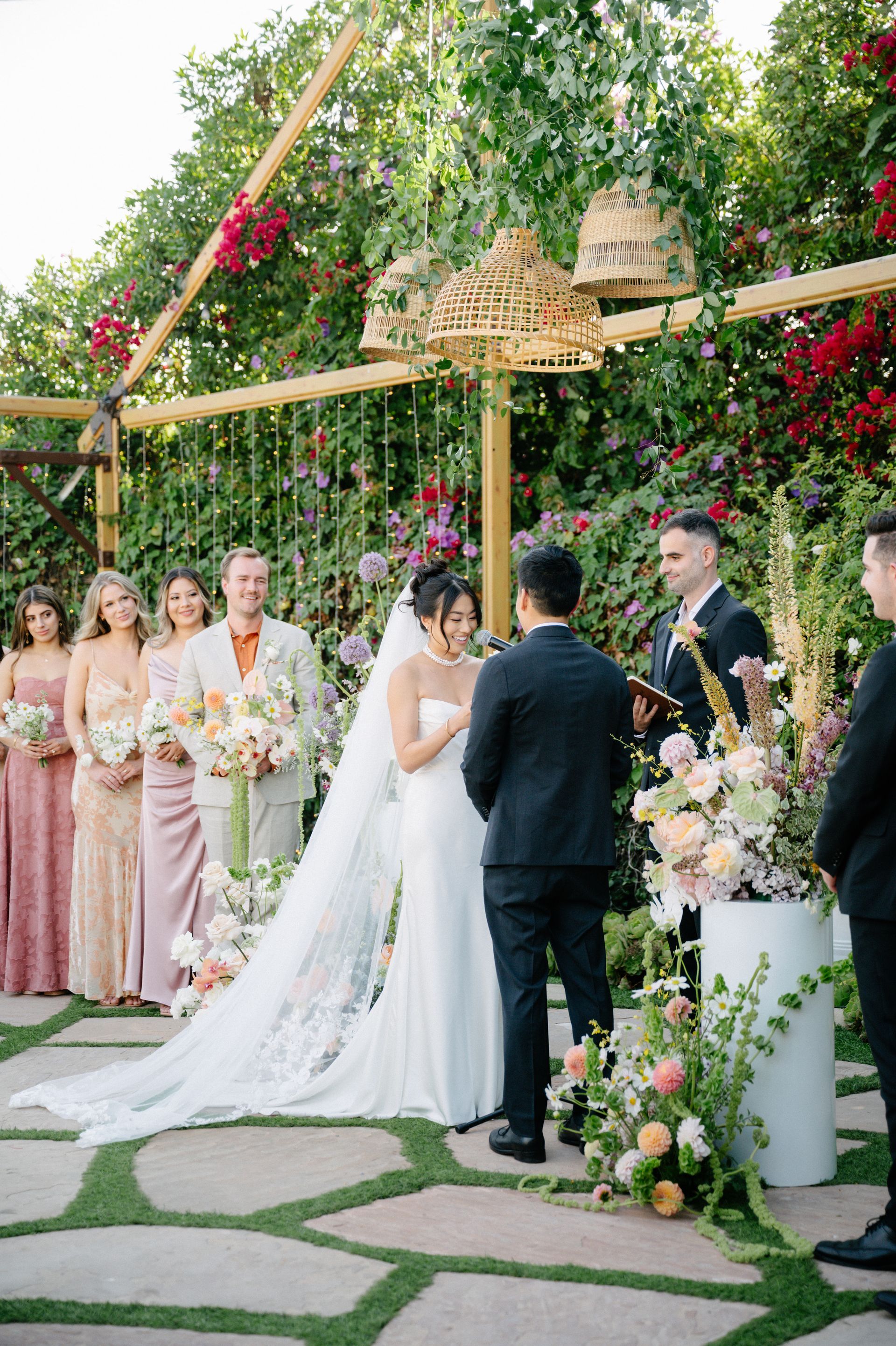 Outdoor wedding ceremony with bride and groom under floral arch, bridesmaids and guests nearby