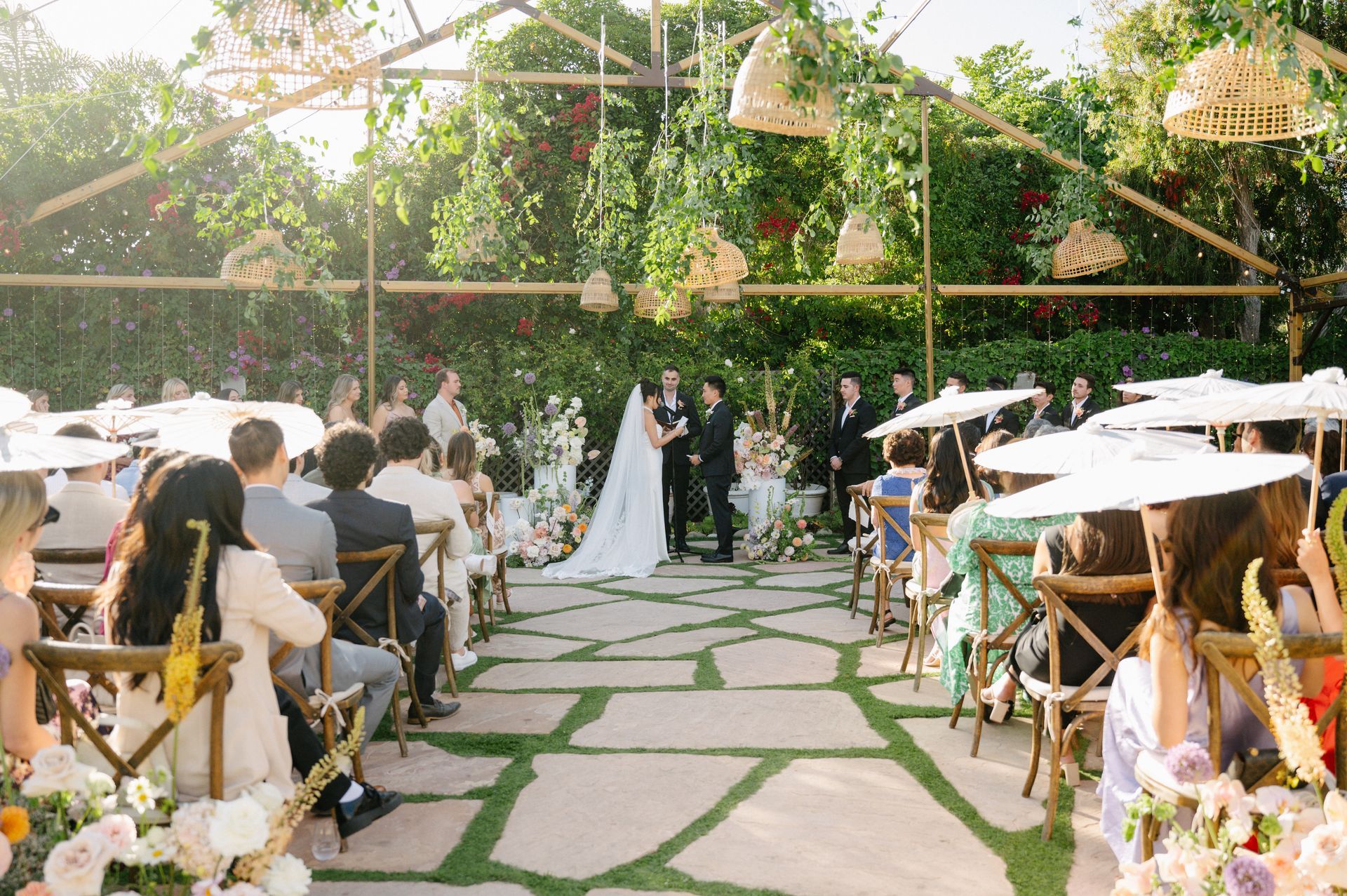 Outdoor wedding ceremony with white aisle chairs, stone path, and bride and groom under greenery