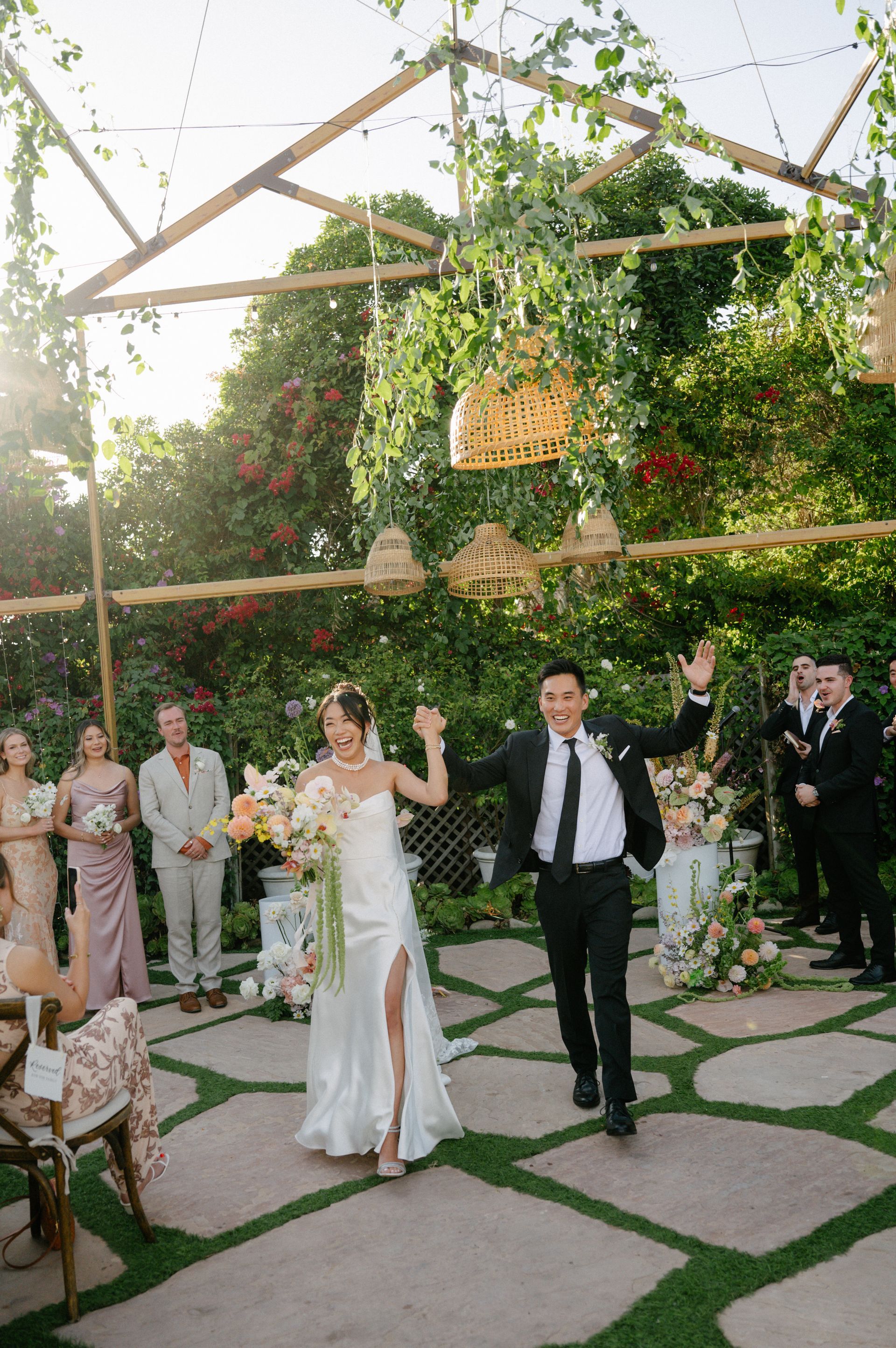 Wedding couple walking down a vine-covered aisle outdoors, guests applauding around them