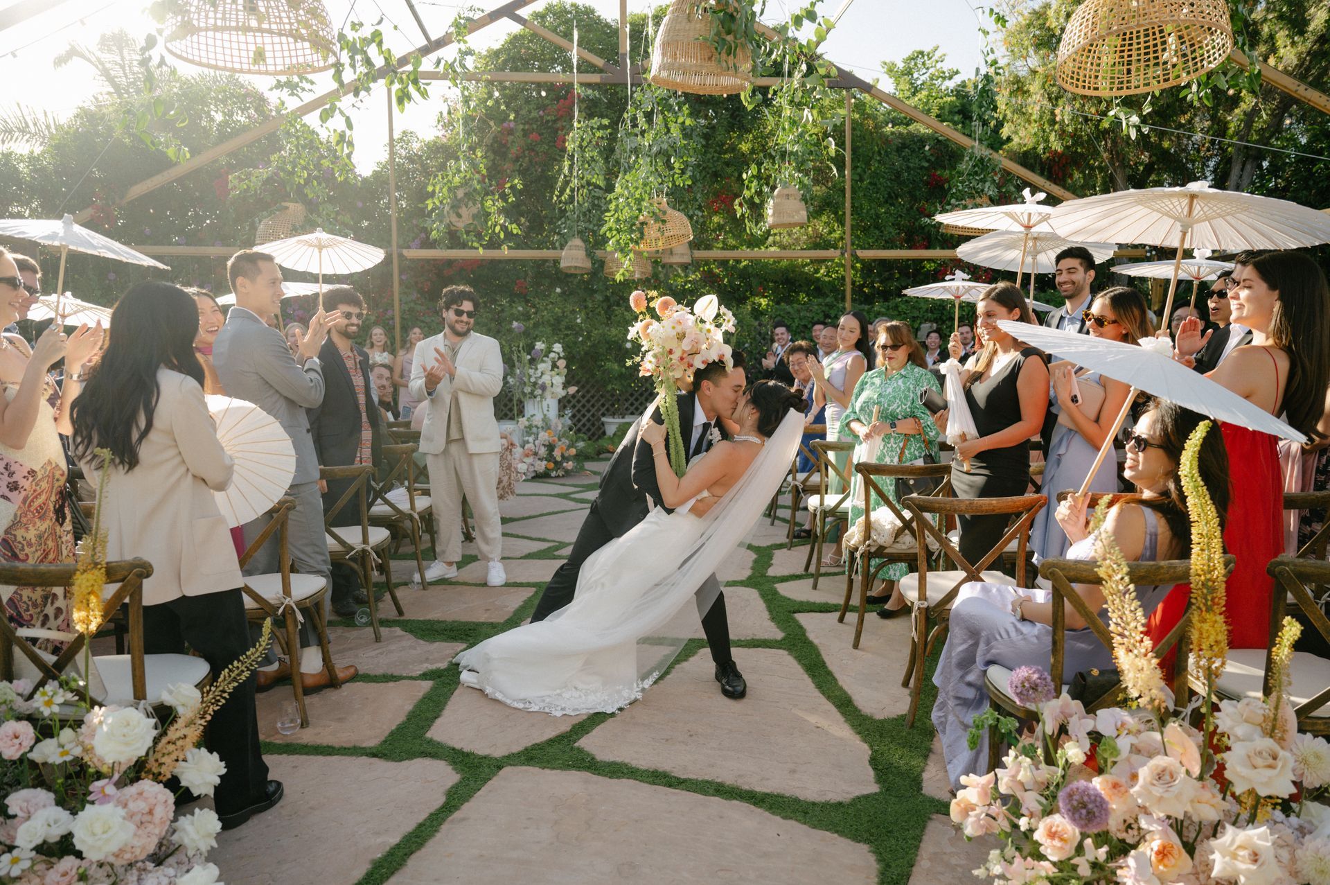 Outdoor wedding ceremony with a bride in white walking down a flower-lined aisle under a pergola
