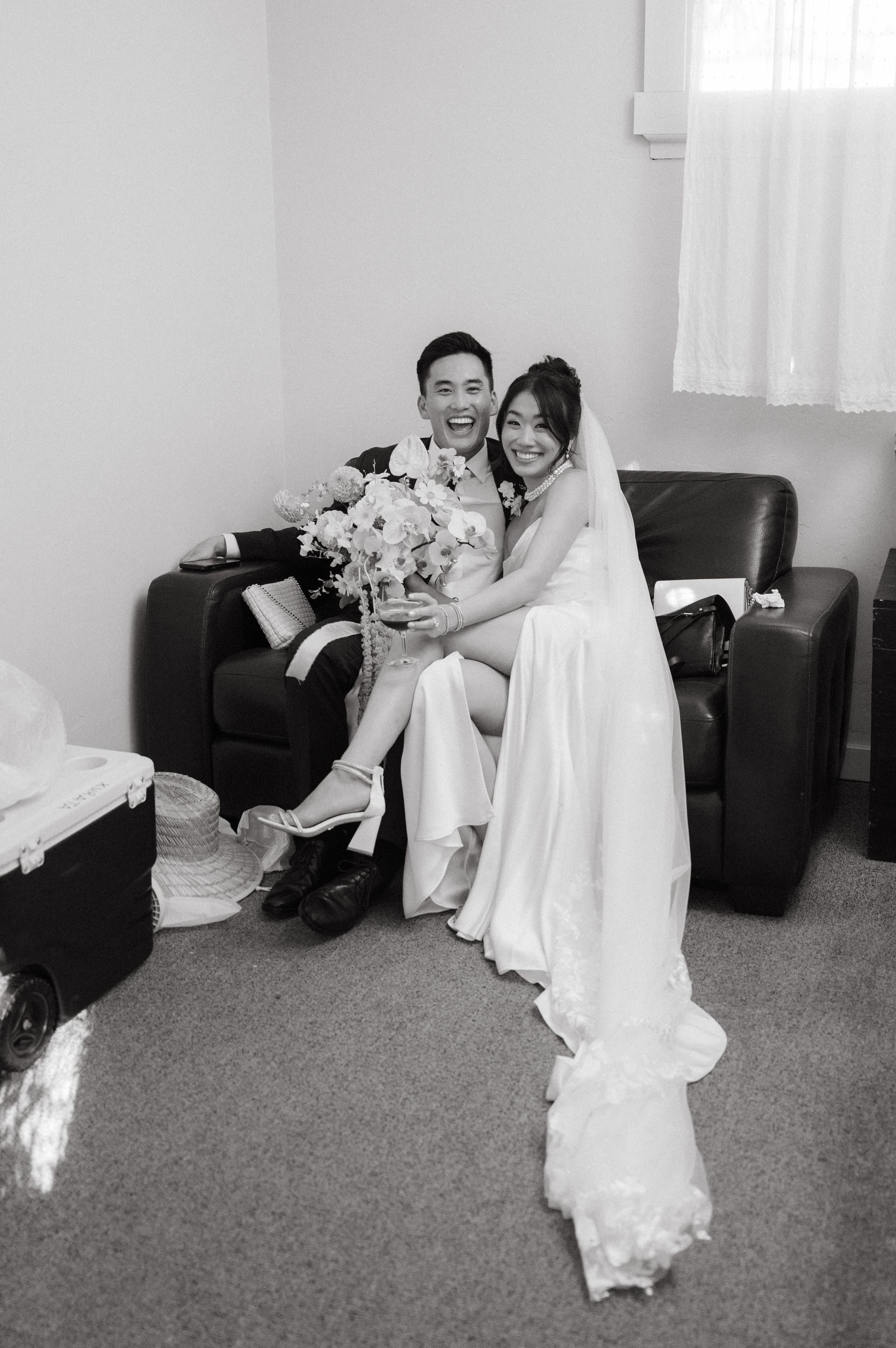 Bride and groom seated on a couch, smiling and holding a bouquet in a small room.