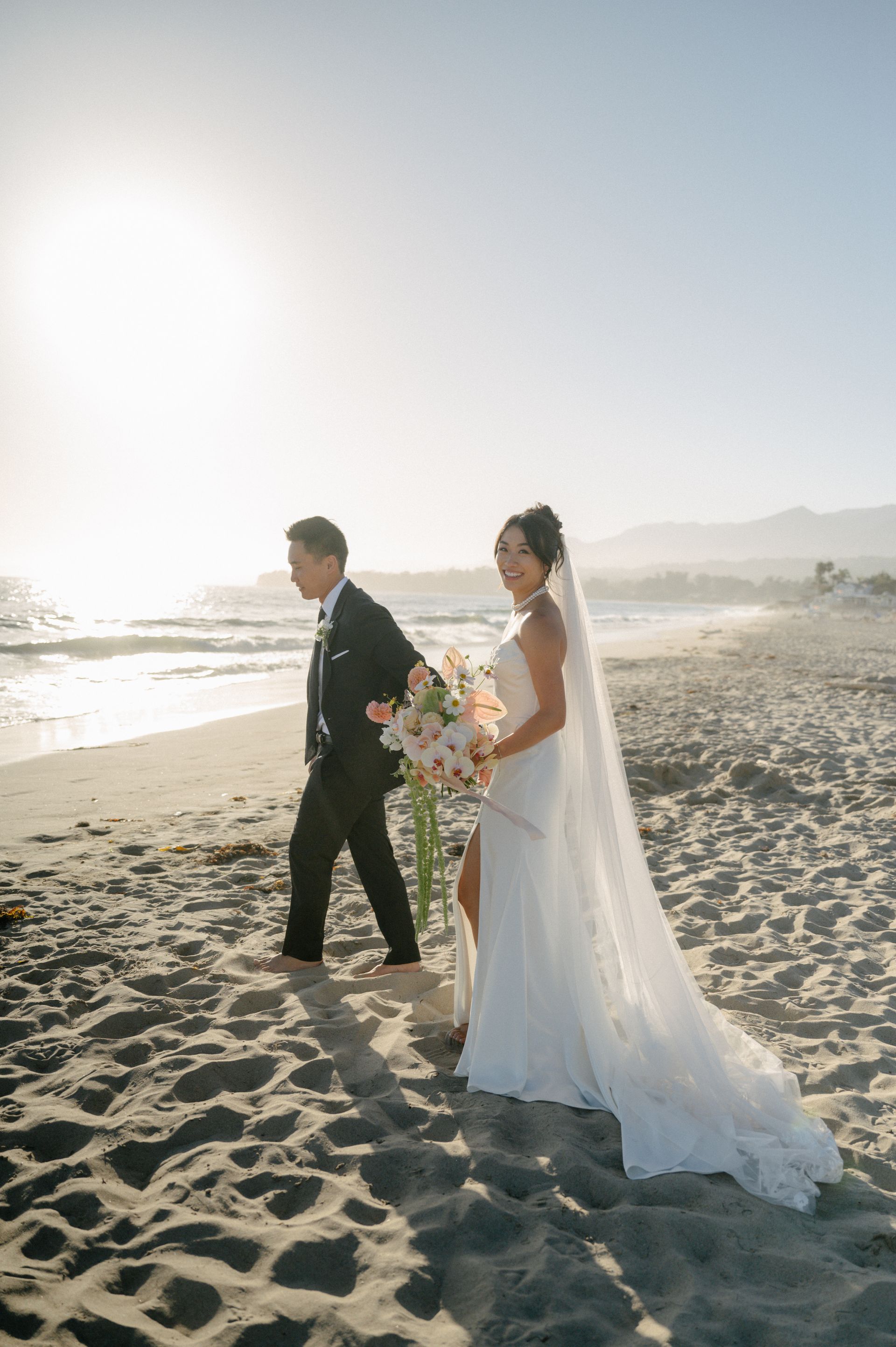Bride and groom walking on a sunny beach, holding hands with a bouquet, dress trailing in the sand