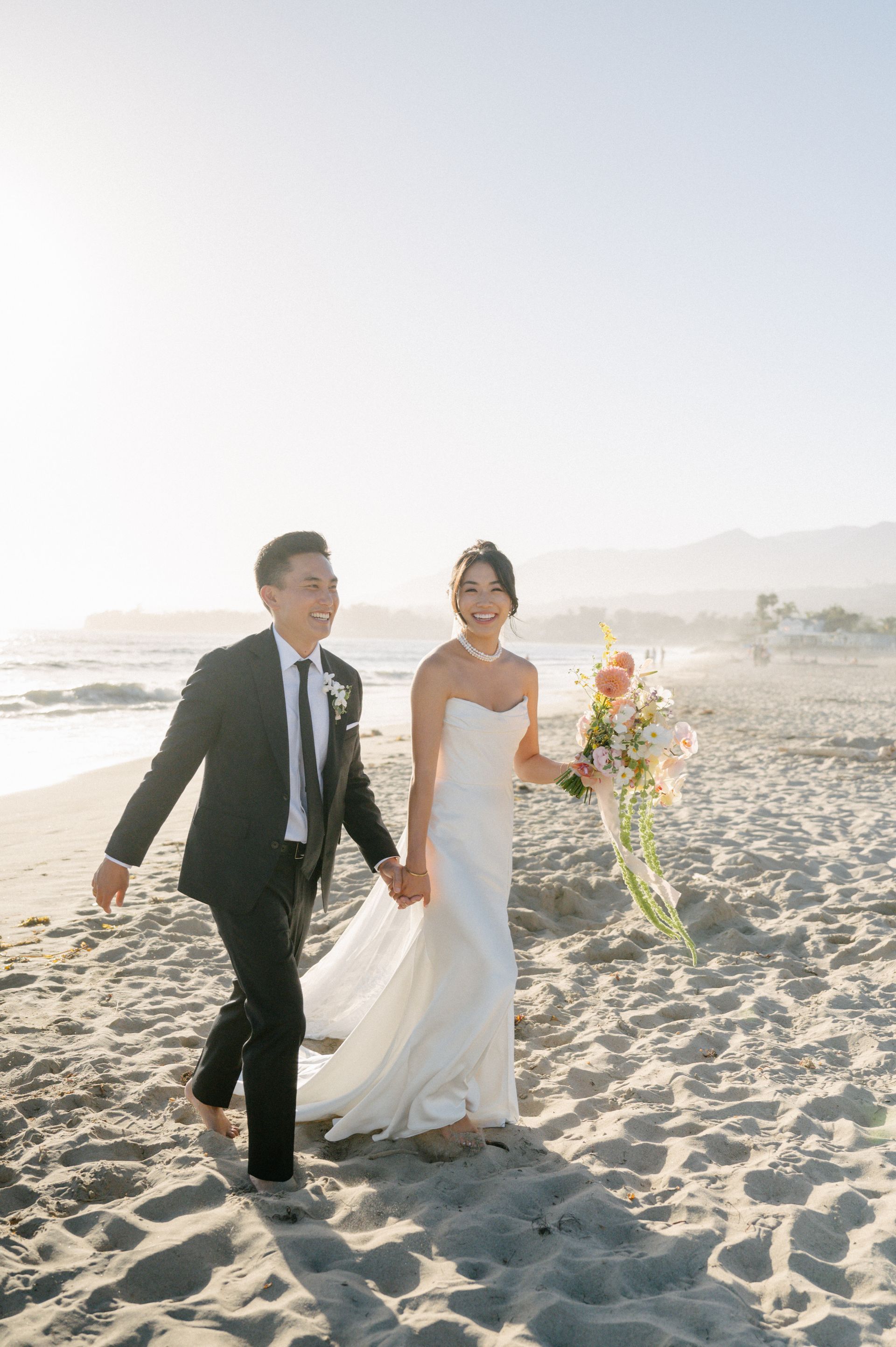 Bride and groom walking hand in hand on a sunny beach, bride holding a bouquet.
