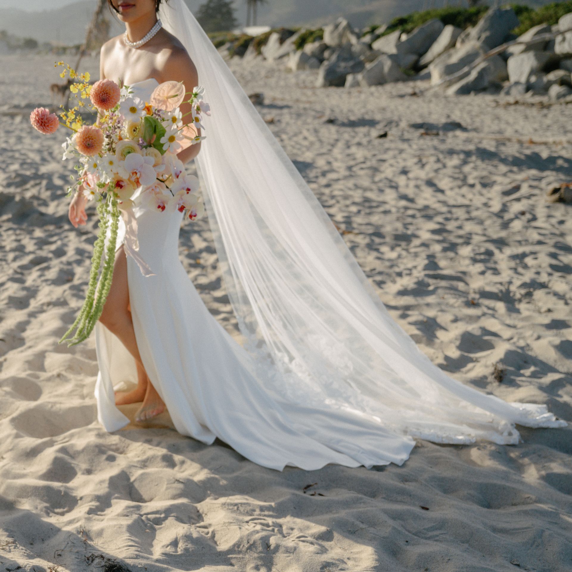 Bride in a long white dress holding a colorful bouquet on a sandy beach with a flowing veil
