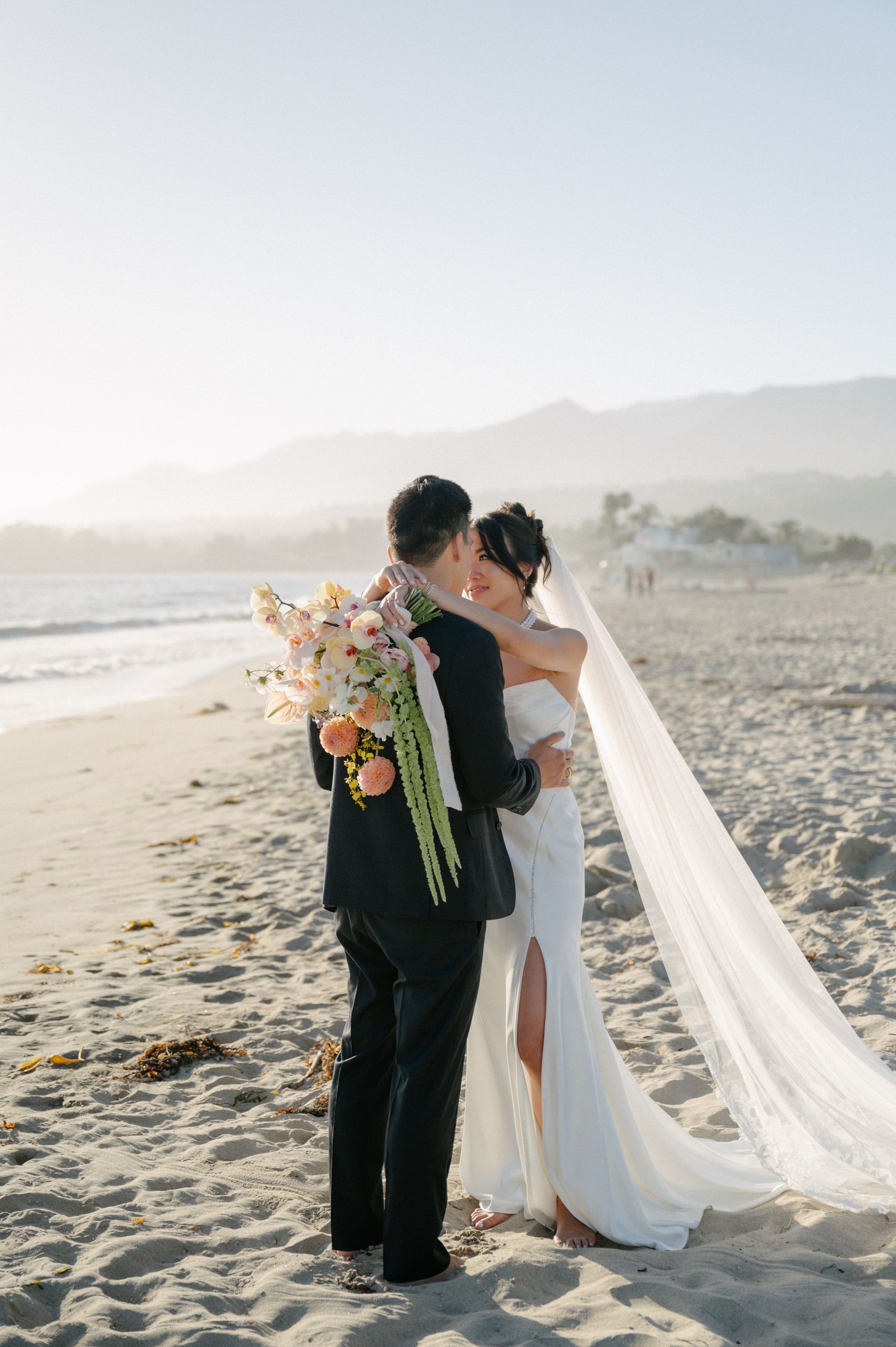 Bride and groom embracing on a beach at sunset, with a long white veil flowing behind her.