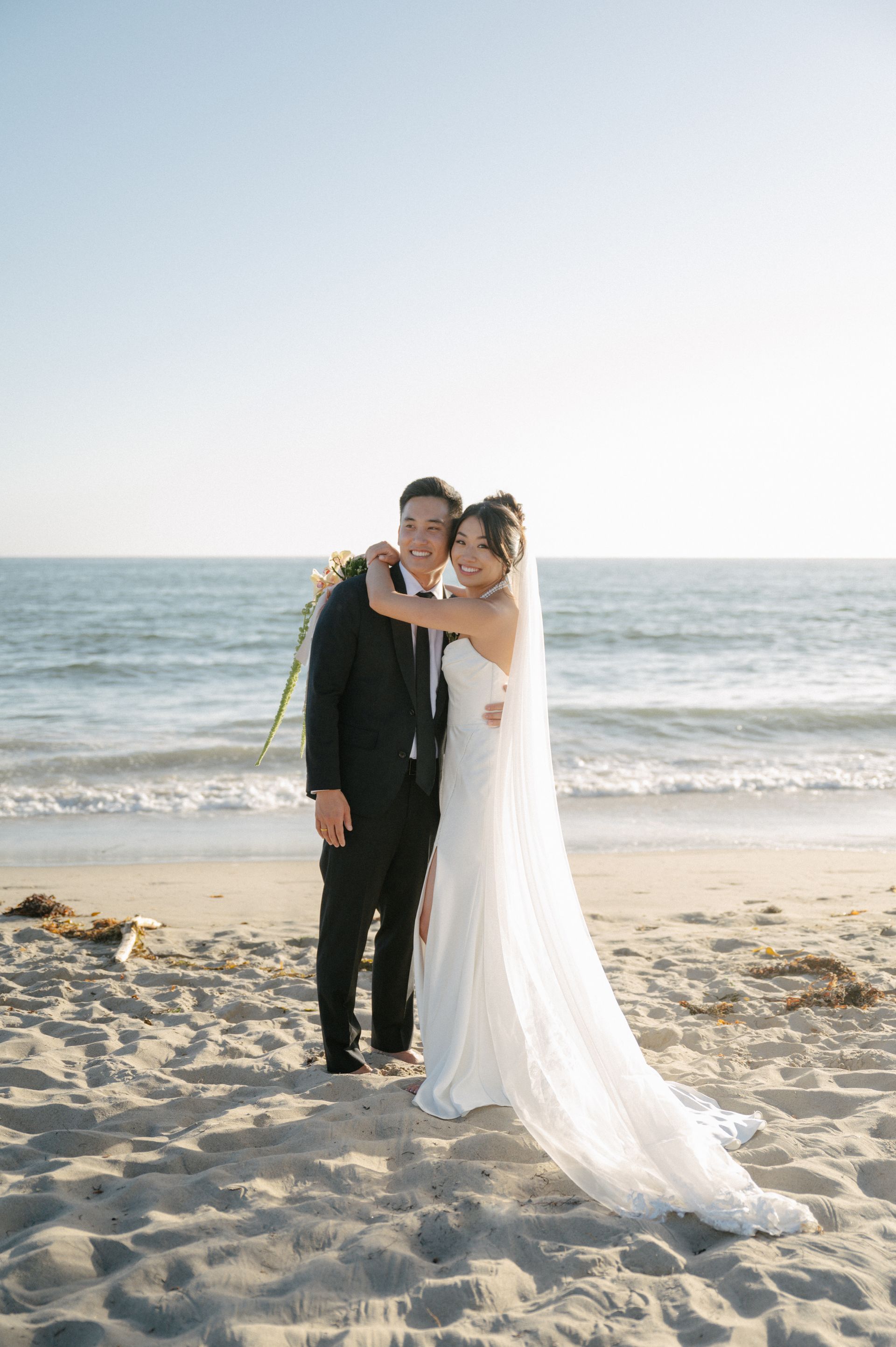 Bride and groom embracing on a beach at sunset, with waves and sand in the background